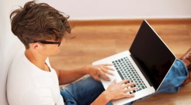 Young man typing on a laptop while sitting on a wooden floor in a bright room, wearing a white T‑shirt and jeans.