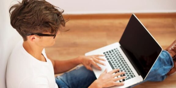 Young man typing on a laptop while sitting on a wooden floor in a bright room, wearing a white T‑shirt and jeans.