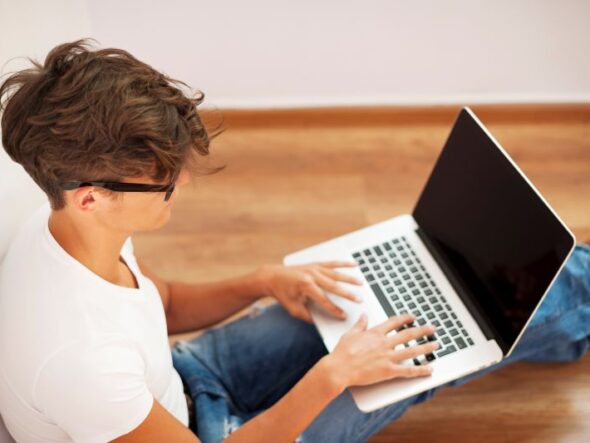 Young man typing on a laptop while sitting on a wooden floor in a bright room, wearing a white T‑shirt and jeans.