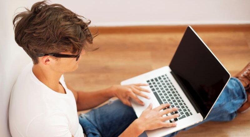 Young man typing on a laptop while sitting on a wooden floor in a bright room, wearing a white T‑shirt and jeans.
