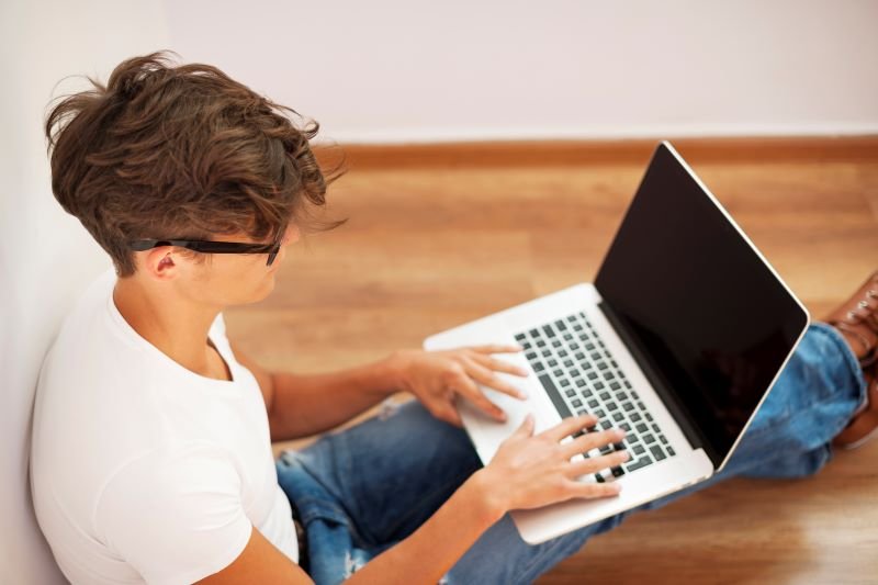 Young man typing on a laptop while sitting on a wooden floor in a bright room, wearing a white T‑shirt and jeans.