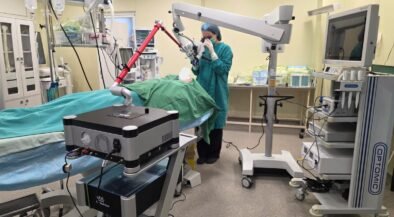 Surgeon in teal scrubs operates a robotic-assisted system over a patient on an operating table in a bright OR.