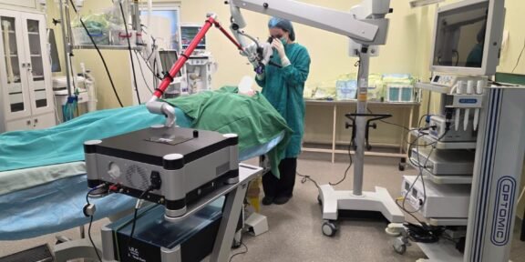 Surgeon in teal scrubs operates a robotic-assisted system over a patient on an operating table in a bright OR.