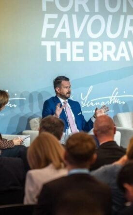 Panel discussion with three speakers on stage; backdrop reads 'Fortune Favours The Brave'.