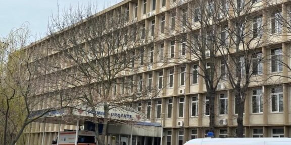 Exterior view of a multi-story hospital building with leafless trees and an ambulance parked at the urgent care entrance.
