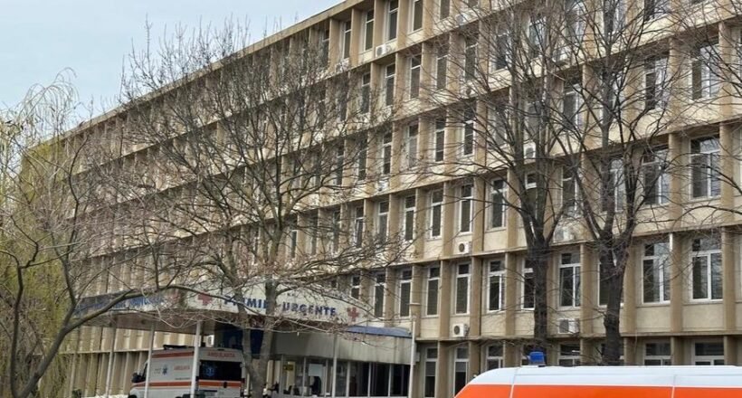Exterior view of a multi-story hospital building with leafless trees and an ambulance parked at the urgent care entrance.
