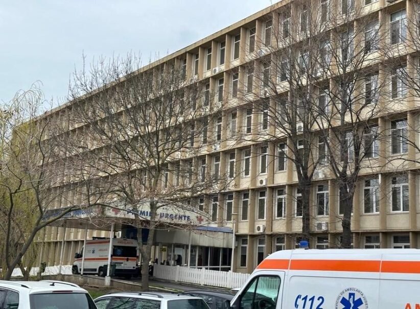Exterior view of a multi-story hospital building with leafless trees and an ambulance parked at the urgent care entrance.