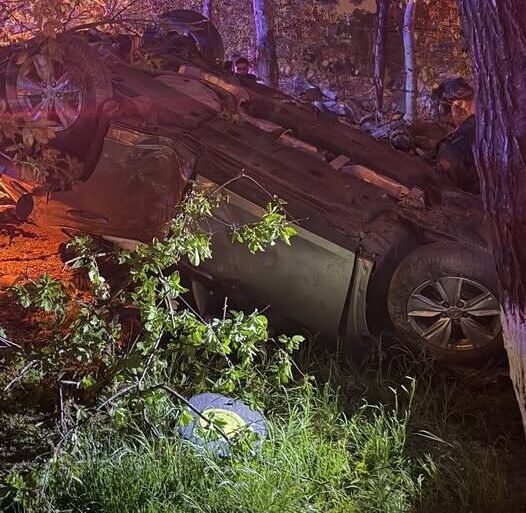 Upside-down car crashed in a wooded area at night with trees and grass around, purple emergency lights nearby.