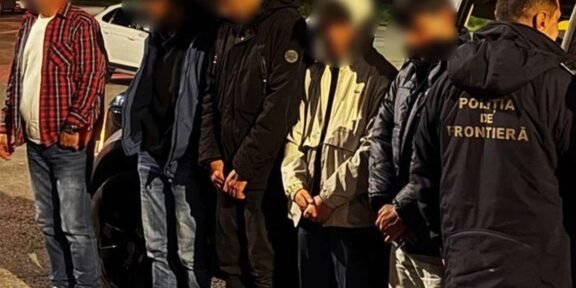 Detainees with hands bound stand in a line at night beside a border police officer wearing a jacket that reads 'Poliția de Frontieră'.
