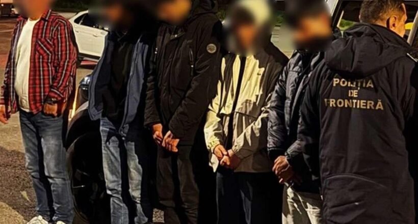 Detainees with hands bound stand in a line at night beside a border police officer wearing a jacket that reads 'Poliția de Frontieră'.