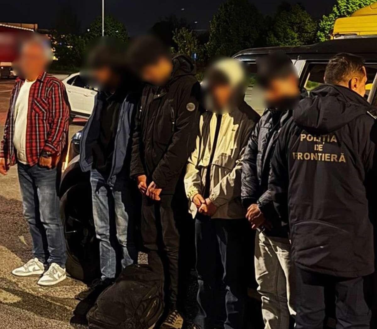 Detainees with hands bound stand in a line at night beside a border police officer wearing a jacket that reads 'Poliția de Frontieră'.