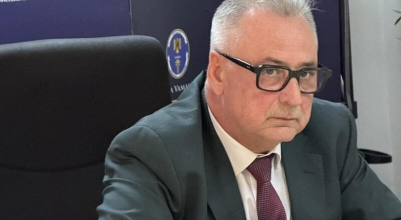 Businessman in a dark suit and maroon tie sits at a desk, wearing black glasses, in a professional office with customs signage in the background.