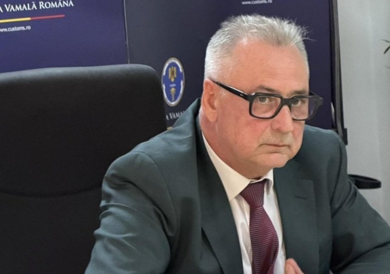 Businessman in a dark suit and maroon tie sits at a desk, wearing black glasses, in a professional office with customs signage in the background.
