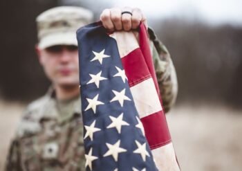 Soldier in camouflage holds an American flag toward the camera in an outdoor, blurred setting behind him.