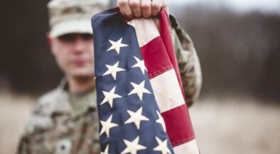 Soldier in camouflage holds an American flag toward the camera in an outdoor, blurred setting behind him.