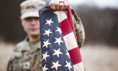 Soldier in camouflage holds an American flag toward the camera in an outdoor, blurred setting behind him.