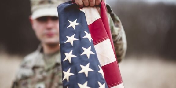 Soldier in camouflage holds an American flag toward the camera in an outdoor, blurred setting behind him.