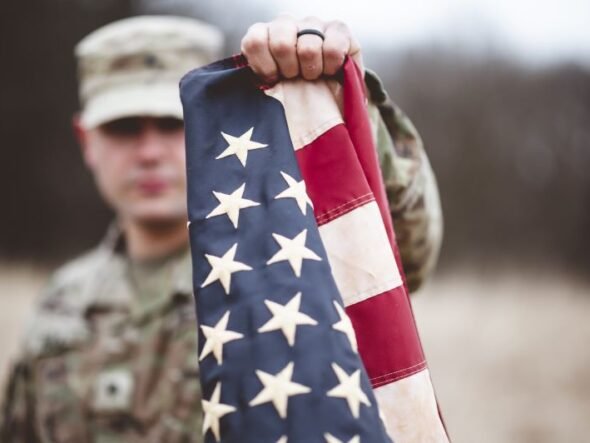 Soldier in camouflage holds an American flag toward the camera in an outdoor, blurred setting behind him.