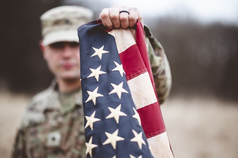Soldier in camouflage holds an American flag toward the camera in an outdoor, blurred setting behind him.