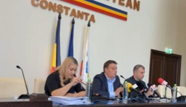 Press conference with three officials seated at a long desk, microphones in front of them, flags behind, and a Constanța signage.