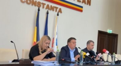 Press conference with three officials seated at a long desk, microphones in front of them, flags behind, and a Constanța signage.