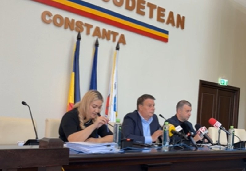 Press conference with three officials seated at a long desk, microphones in front of them, flags behind, and a Constanța signage.