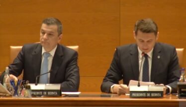 Two men in dark suits sit at a wood-paneled conference table with microphones and nameplates during a formal meeting. Left man looks ahead while right man reads notes into a microphone.