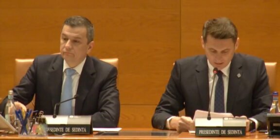 Two men in dark suits sit at a wood-paneled conference table with microphones and nameplates during a formal meeting. Left man looks ahead while right man reads notes into a microphone.