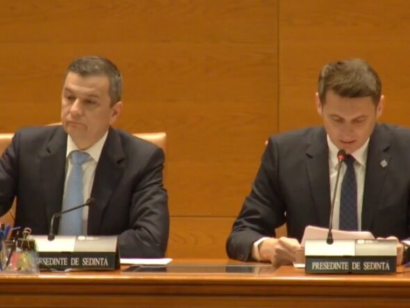 Two men in dark suits sit at a wood-paneled conference table with microphones and nameplates during a formal meeting. Left man looks ahead while right man reads notes into a microphone.