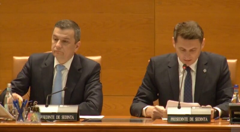 Two men in dark suits sit at a wood-paneled conference table with microphones and nameplates during a formal meeting. Left man looks ahead while right man reads notes into a microphone.