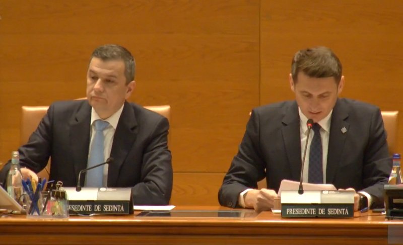 Two men in dark suits sit at a wood-paneled conference table with microphones and nameplates during a formal meeting. Left man looks ahead while right man reads notes into a microphone.
