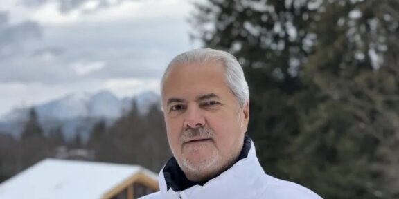 Older man with gray hair wearing a white winter jacket, posing outdoors in a snowy landscape with trees and mountains behind him.