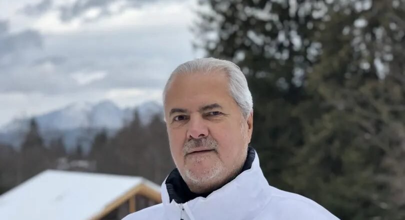 Older man with gray hair wearing a white winter jacket, posing outdoors in a snowy landscape with trees and mountains behind him.