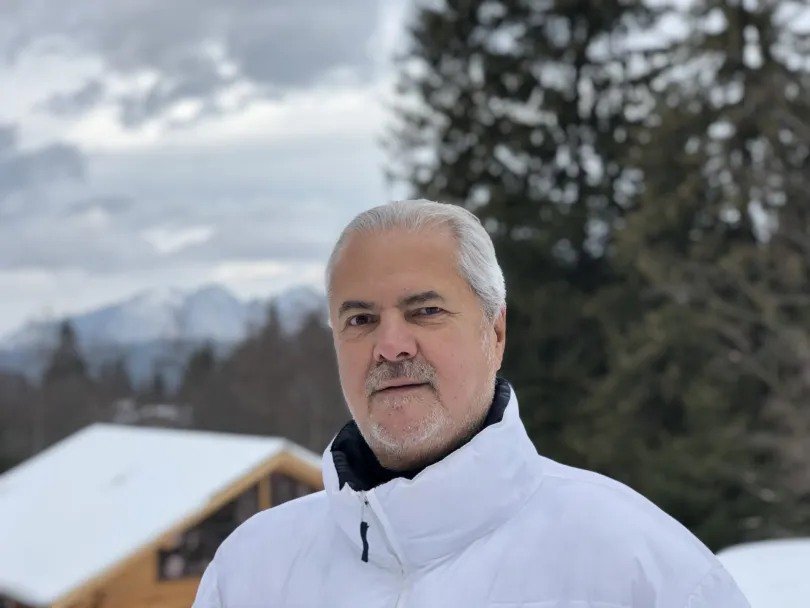 Older man with gray hair wearing a white winter jacket, posing outdoors in a snowy landscape with trees and mountains behind him.