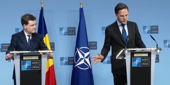 Two suited men stand at podiums during a NATO press conference, with a Romanian flag and NATO flag between them behind a blue backdrop marked NATO OTAN.