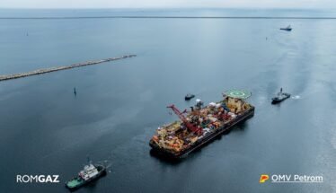 Offshore support vessel with crane and helipad, being moved by two tugboats in open sea; ROMGAZ and OMV Petrom logos visible.