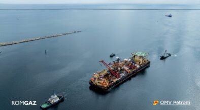 Offshore support vessel with crane and helipad, being moved by two tugboats in open sea; ROMGAZ and OMV Petrom logos visible.