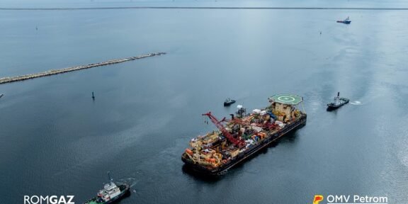 Offshore support vessel with crane and helipad, being moved by two tugboats in open sea; ROMGAZ and OMV Petrom logos visible.
