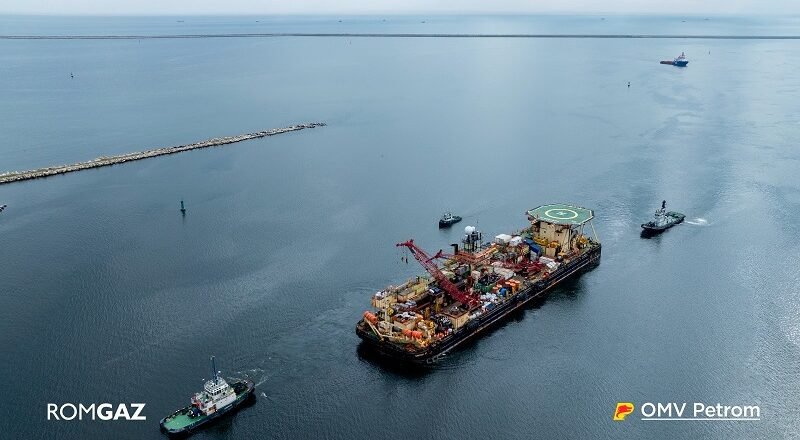 Offshore support vessel with crane and helipad, being moved by two tugboats in open sea; ROMGAZ and OMV Petrom logos visible.