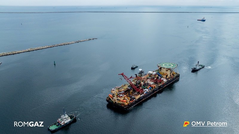 Offshore support vessel with crane and helipad, being moved by two tugboats in open sea; ROMGAZ and OMV Petrom logos visible.