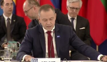 Man in a dark suit and red tie speaks into a microphone at a formal meeting, with colorful flags in the background.