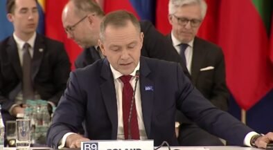 Man in a dark suit and red tie speaks into a microphone at a formal meeting, with colorful flags in the background.