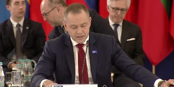 Man in a dark suit and red tie speaks into a microphone at a formal meeting, with colorful flags in the background.