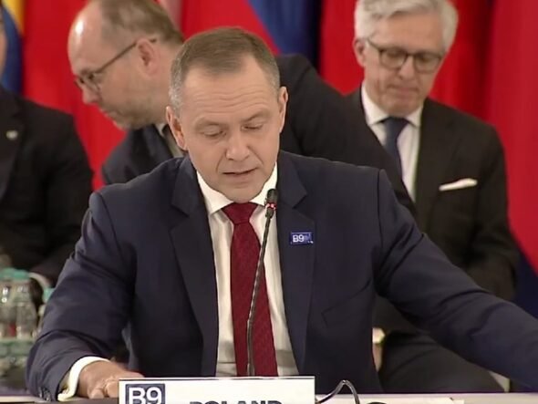 Man in a dark suit and red tie speaks into a microphone at a formal meeting, with colorful flags in the background.