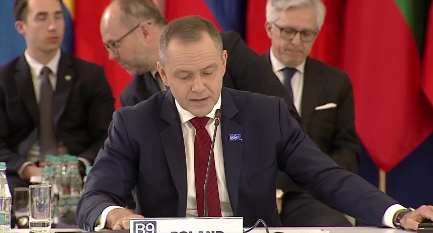 Man in a dark suit and red tie speaks into a microphone at a formal meeting, with colorful flags in the background.