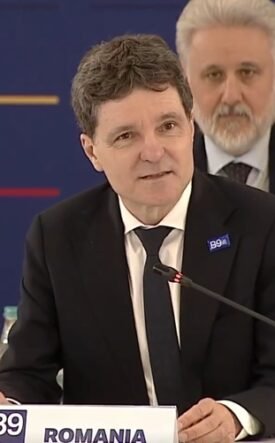 Man in a dark suit and tie speaking at a conference podium labeled 'ROMANIA' with a microphone and seated officials in the background.