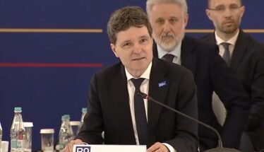 Man in a dark suit and tie speaking at a conference podium labeled 'ROMANIA' with a microphone and seated officials in the background.