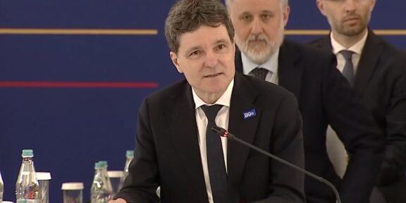 Man in a dark suit and tie speaking at a conference podium labeled 'ROMANIA' with a microphone and seated officials in the background.