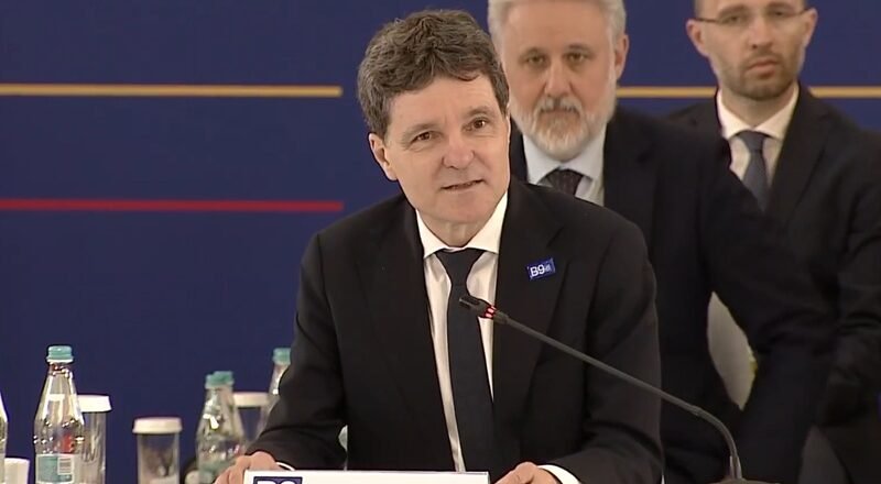 Man in a dark suit and tie speaking at a conference podium labeled 'ROMANIA' with a microphone and seated officials in the background.