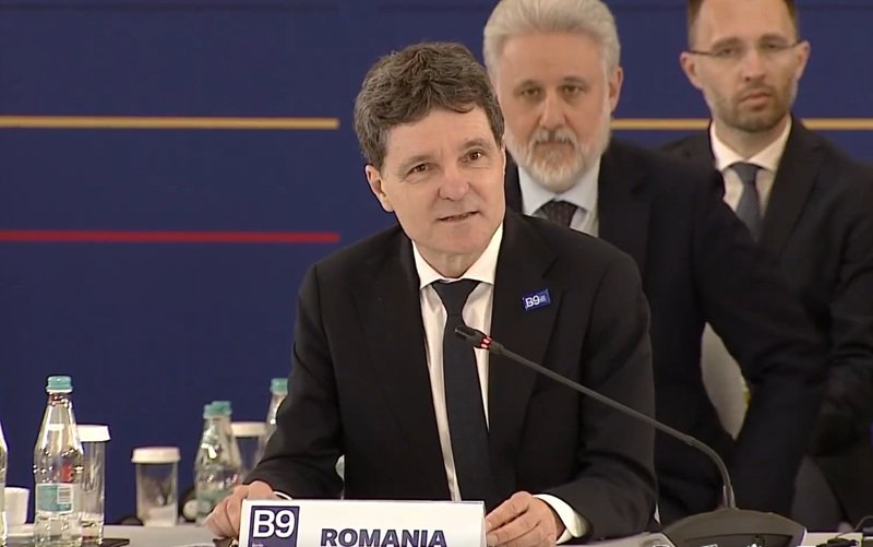 Man in a dark suit and tie speaking at a conference podium labeled 'ROMANIA' with a microphone and seated officials in the background.